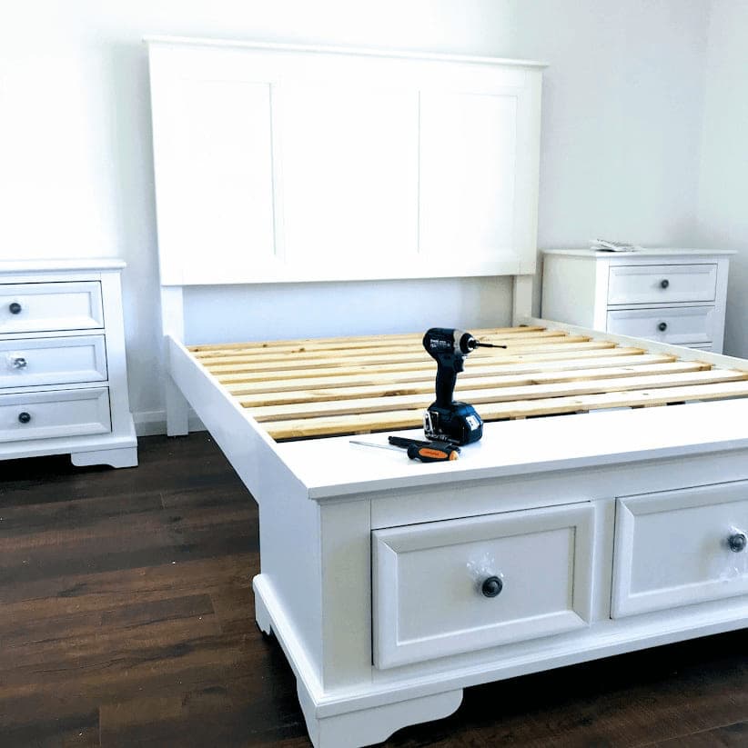White panelled bed frame with a timber slat base and end-of-bed storage drawers, part-assembled between two matching bedside tables with the drill still on the footboard