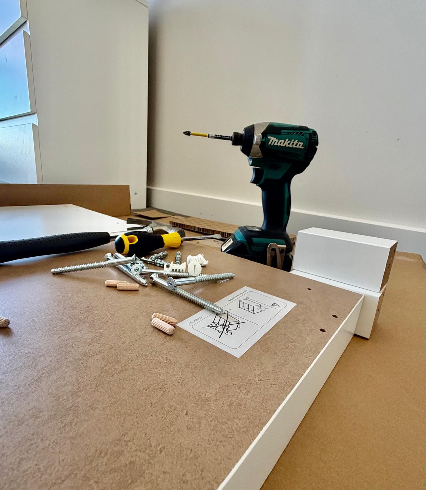 Flat-pack panel drilled for cam fittings, with dowels, confirmat screws, wall plugs, an instruction sheet, a hammer, a screwdriver and an impact driver laid out mid-assembly