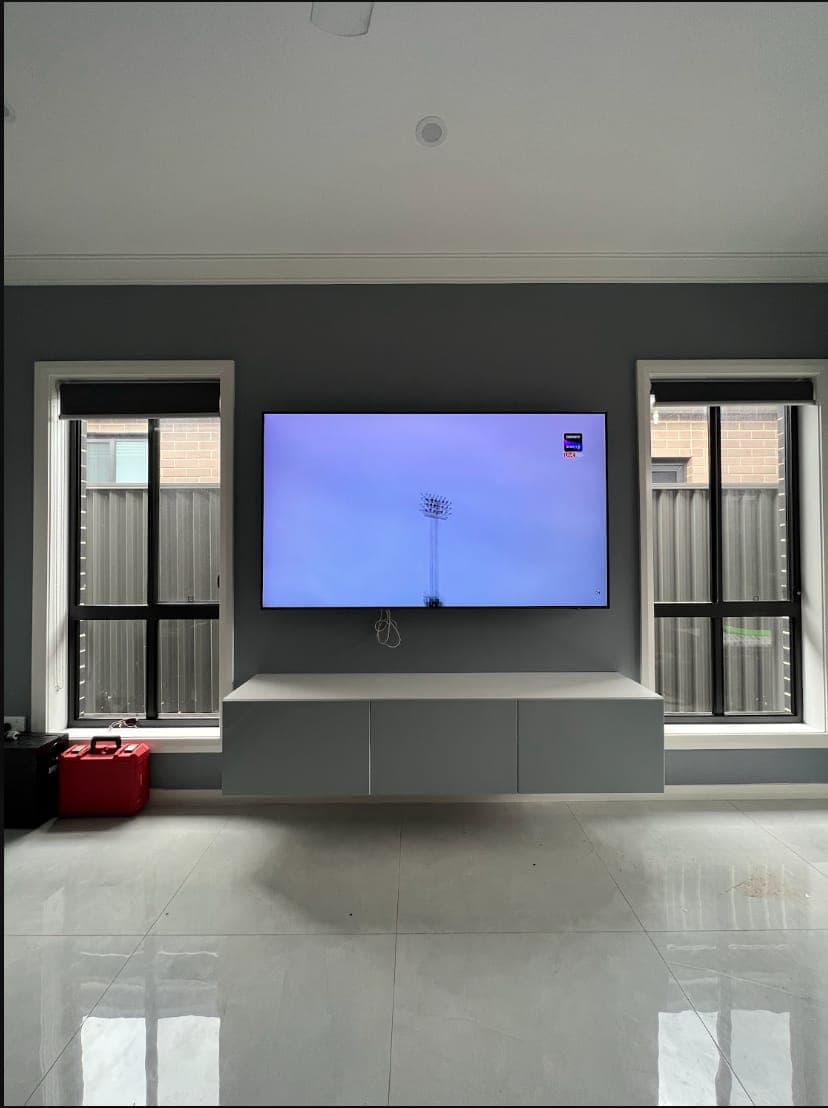 A large flat-screen TV wall mounted on a dark grey feature wall above a white floating cabinet, centred between two windows in a tiled living room