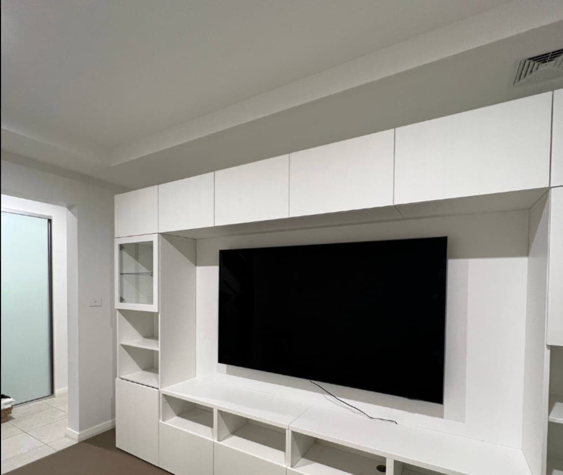 A TV mounted flush inside a white built-in wall unit, framed by overhead cupboards, a glass display cabinet and open shelving