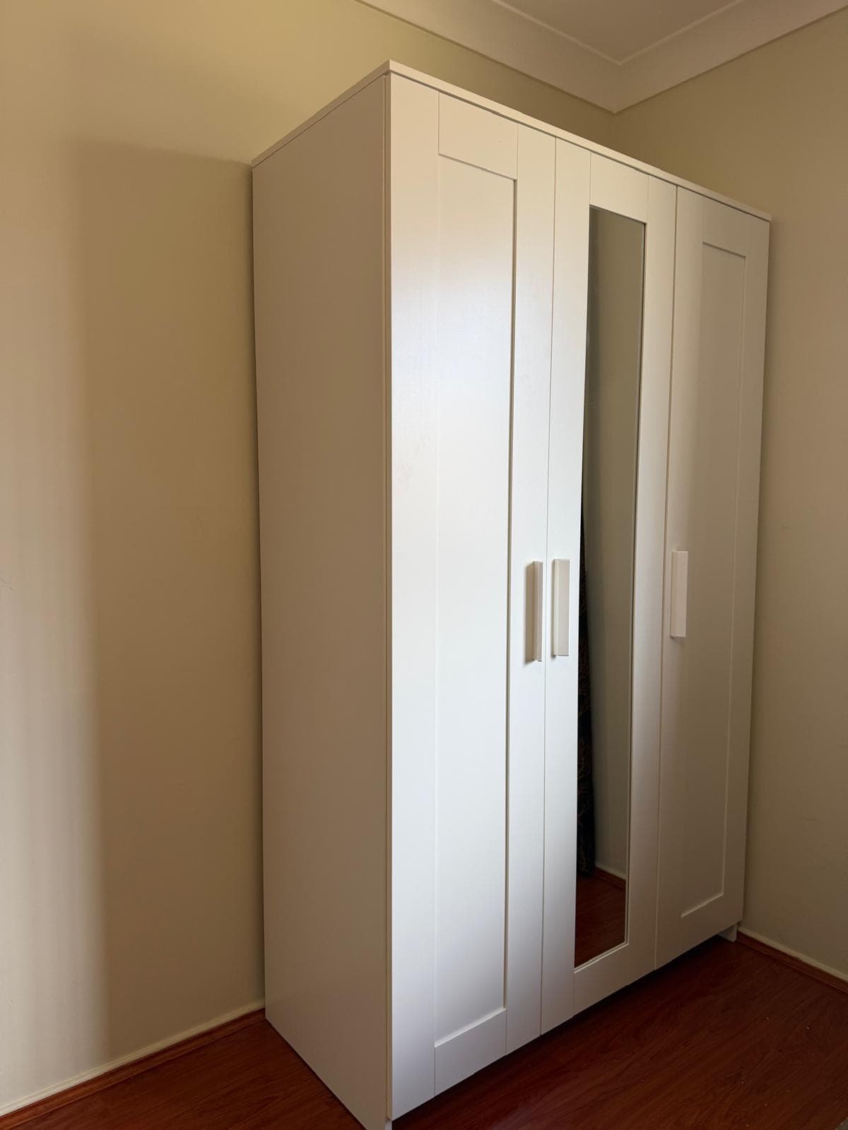 Three-door wardrobe in white with a mirrored centre door and slim vertical handles, assembled and standing against a bedroom wall